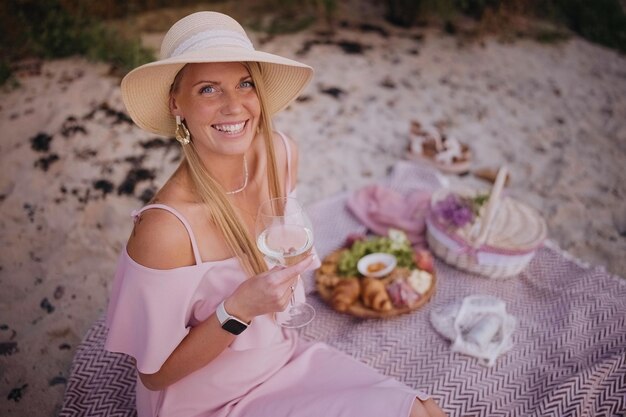 Young woman in straw hat sit on plaid have picnic with wine on sand beach