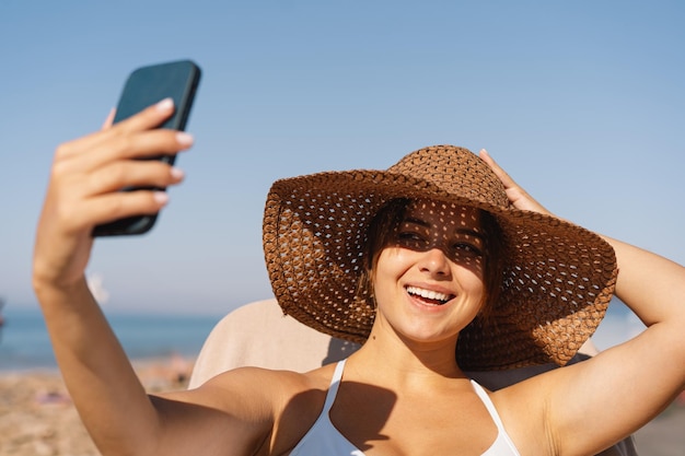 晴れた日にビーチでセルフィーを撮る草の帽子をかぶった若い女性