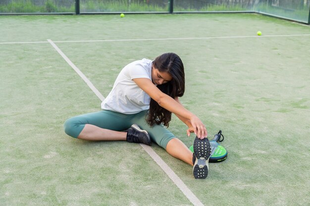 Foto giovane donna che si estende seduta sul pavimento sul campo da padel con la racchetta