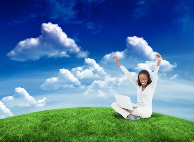 Young woman stretching while on the floor with her laptop against green field under blue sky