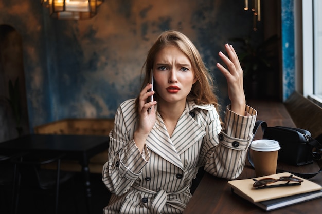 Young woman in striped trench coat emotionally talking on cellphone sadly 