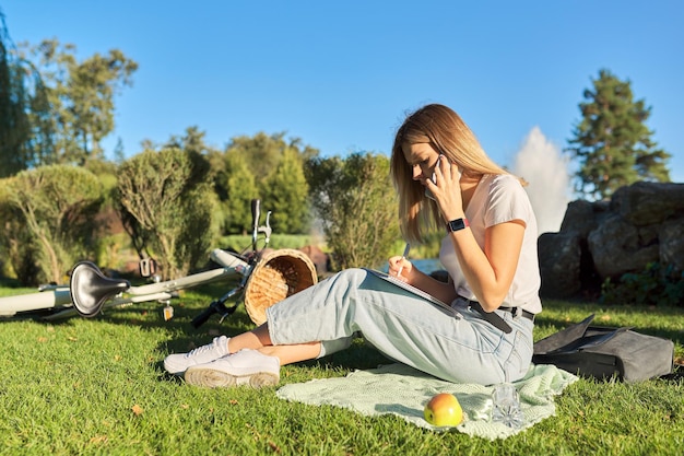 Young woman student 19, 20 years old sitting on grass in park with textbook, girl studies using smartphone, writes in notebook. E-learning, educational technologies, distance learning, self-education