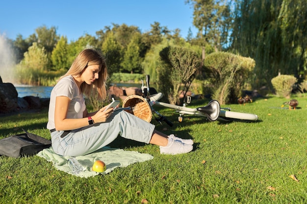 写真 教科書を持って公園の芝生に座っている19、20歳の若い女性学生、スマートフォンを使用して女の子の勉強、ノートに書き込みます。 eラーニング、教育工学、遠隔教育、独学