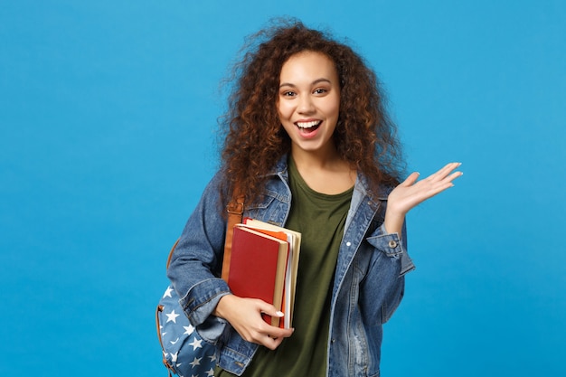 デニムの服とバックパックの若い女性の学生は青い壁に隔離された本を保持します。