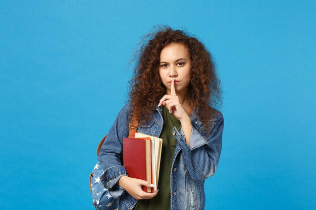 デニムの服とバックパックの若い女性の学生は青い壁に隔離された本を保持します。