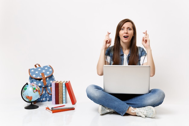 Young woman student hold laptop computer waiting for special moment keeping fingers crossed near globe backpack school books isolated