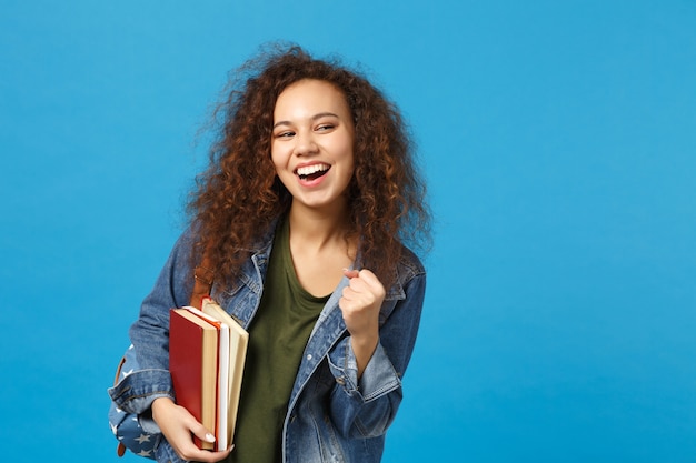 デニムの服とバックパックの若い女性の学生は青い壁に隔離された本を保持します。