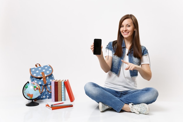 Young woman student pointing index finger on mobile phone with blank black empty screen near globe, backpack, school books isolated on white wall