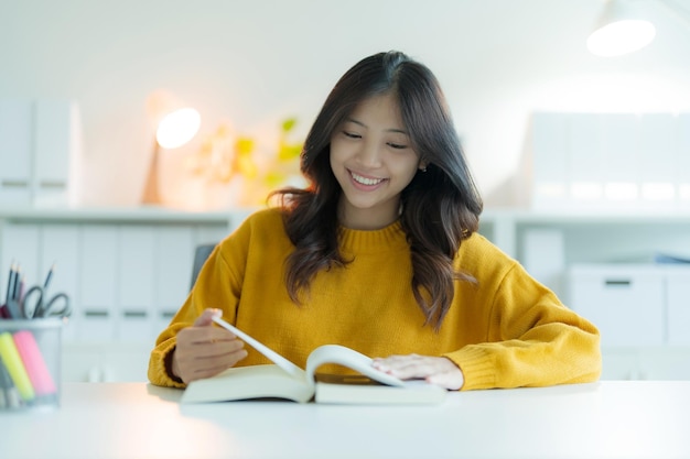 A young woman or student reading a book in the library while wearing glasses She is smiling and looking happy