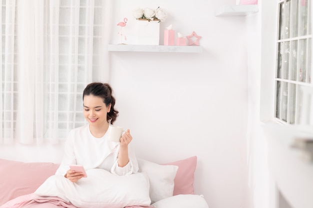 Young woman student sitting on bed in her room drinking coffee and using mobile phone