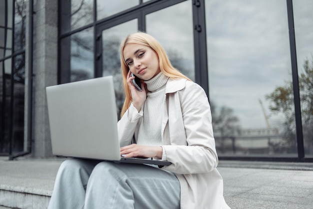 Young woman student sitting with laptop and talking on phone near business center