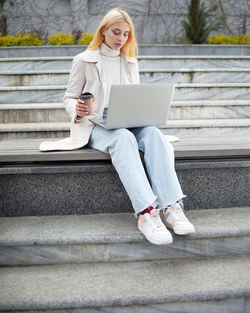 Young woman student uses a laptop and drink coffee while sitting on bench