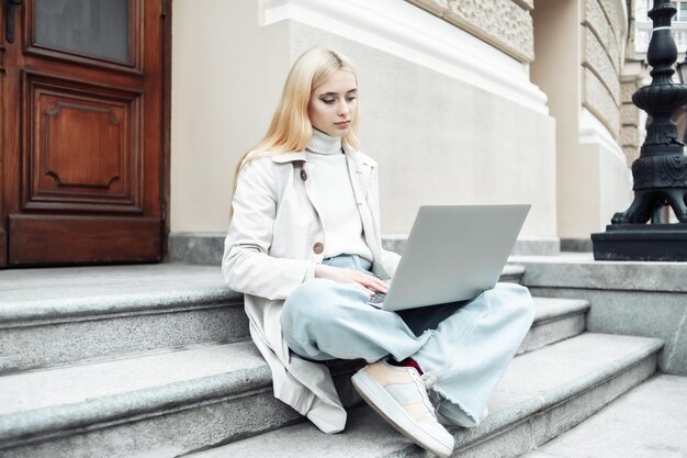 Young woman student uses laptop while sitting on stairs in city