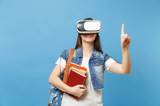 Young woman student in virtual reality headset hold books touch something like push on button, pointing at floating virtual screen isolated on blue background. Education in school university college.