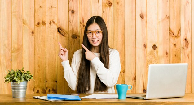 Young woman studying on her desk shocked pointing with index fingers to a copy space