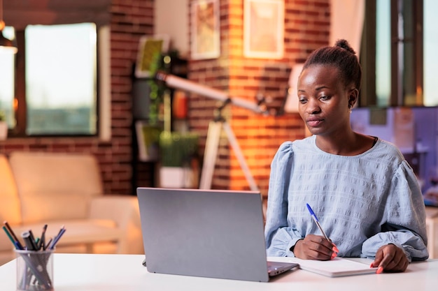 young woman studying, using laptop and taking notes in notepad. female african american remote university student attending remote classes and learning with online courses on internet