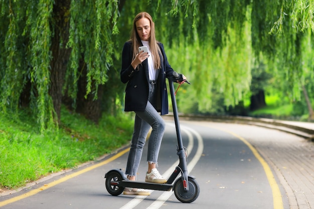 A young woman in stylish clothes poses next to an electric scooter on a bike path