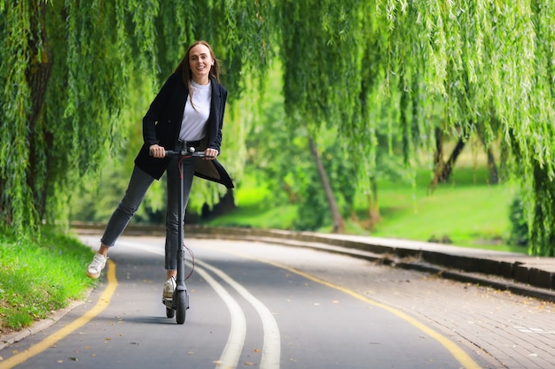 A young woman in stylish clothes rides an electric scooter on a bike path