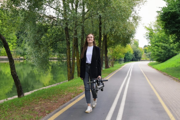 A young woman in stylish clothes walks along a bike path with a folded electric scooter