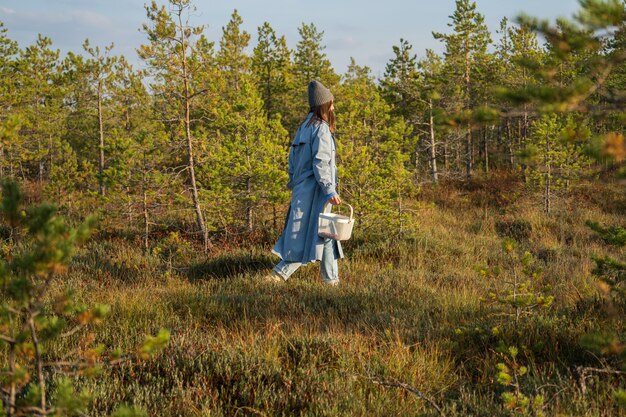 Young woman in stylish coat and hat walk in autumn forest in sunny warm day searching for cranberry