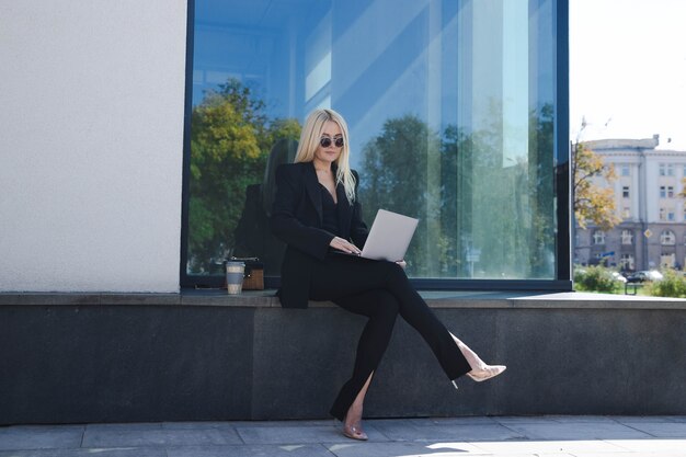 A young woman in a stylish suit is sitting outside and working on a laptop