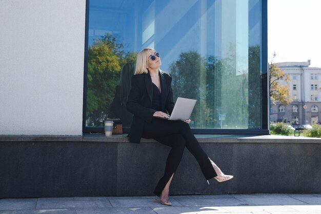 A young woman in a stylish suit is sitting outside and working on a laptop