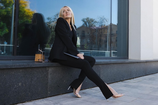 A young woman in a stylish suit is sitting on the street