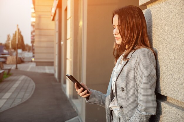 スーツを着た若い女性が建物の近くの通りで電話を見る