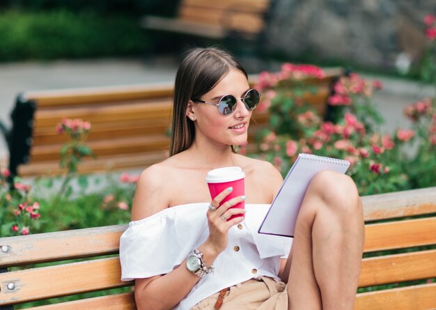 young woman in summer clothes and sunglasses holding cup of coffee in her hands and writing into notebook while sitting on bench in the park