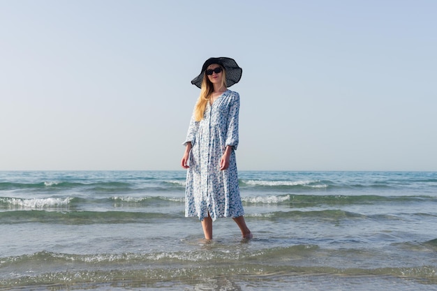 A young woman in a summer dress and hat walks along the seashore