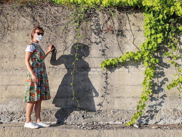 Young woman in summer dress and protective face mask alone\
stands next to concrete wall with ivy