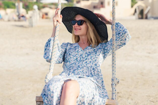 A young woman in a summer dress rides on a swing on the seashore