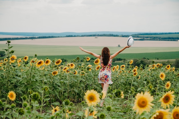 A young woman in a summer dress runs in a field with sunflowers spread her arms to the sides.