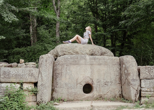 目をそらしている森の大きなドルメン石の上に座っている夏のドレスの若い女性