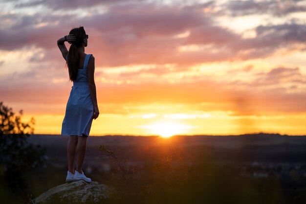 明るい黄色の夕日の景色を楽しみながら屋外に立っている夏のドレスを着た若い女性。