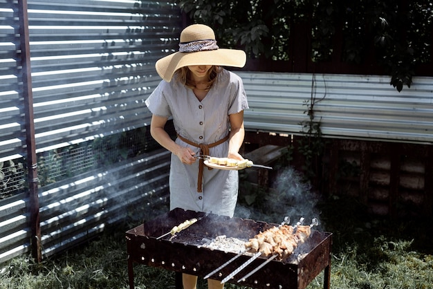 夏の帽子とドレスを着た若い女性が、裏庭で屋外で肉や野菜を焼いています。