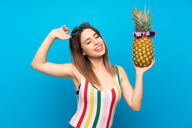 Young woman in summer holidays over blue background holding a pineapple with sunglasses