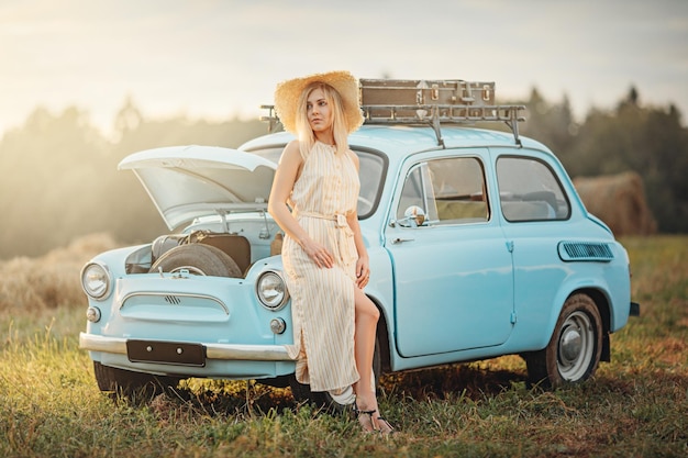 Young woman in sundress and straw hat standing near a blue vintage car with suitcases on a roof rack and opened hood waitng for help summer vibe