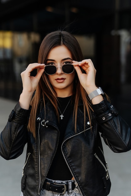 Young woman in sunglasses, hat and black leather jacket posing outdoor.