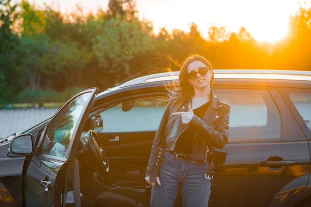 日没の黒い車の近くにサングラスをかけた若い女性