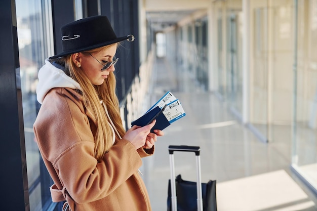 サングラス、暖かい服と帽子の若い女性が手に電話とチケットを持って空港の屋内に立っています。