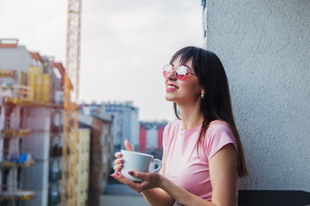 一杯のコーヒーとサングラスの若い女性はバルコニーに滞在します。