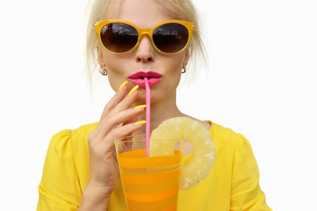 A young woman in sunglasses and a yellow dress drinks juice from a straw.