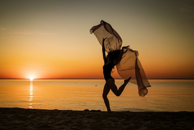 Young woman at sunset jumping on the seashore with a cloth on the background of the sky