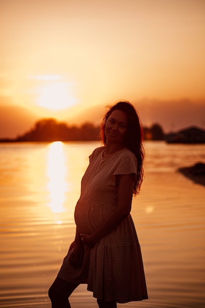 Young woman at sunset silhouette of pregnant happy woman with long hair in rays of setting sun stand...