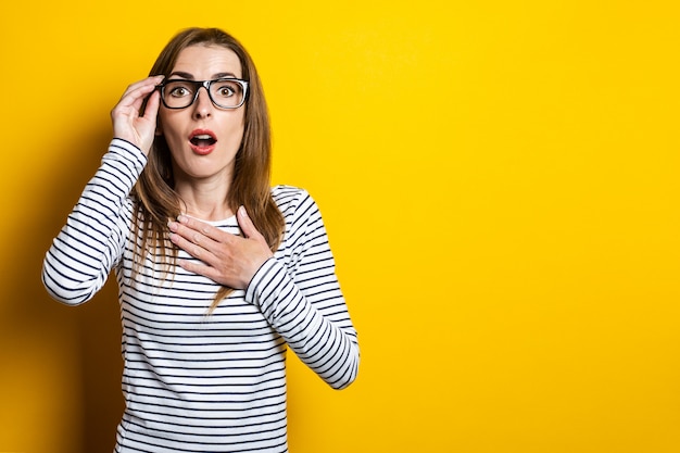 Young woman surprised in shock holding glasses on a yellow background
