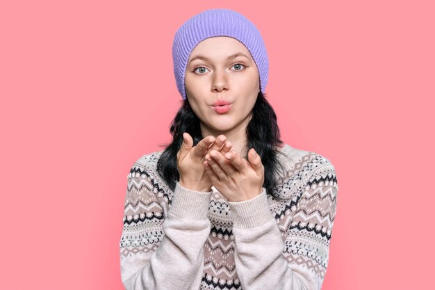 Young woman in sweater hat blowing air kiss on pink color background