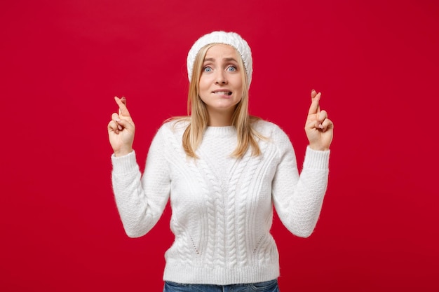 Photo young woman in sweater, hat isolated on red background in studio. healthy fashion lifestyle cold season concept. mock up copy space. waiting for special moment, keeping fingers crossed, making wish.