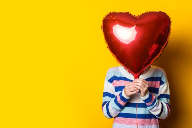 Young woman in a sweater hides behind an air balloon heart on a yellow background. Valentine's day composition.