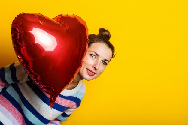 Young woman in a sweater hides behind an air balloon heart on a yellow background. Valentine's day composition.